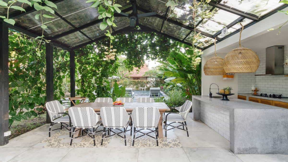 Een terrasoverkapping naast een buitenkeuken met glazen dak en aluminium of stalen profielen, begroeid met planten.