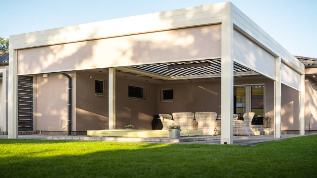 Een grote beige-wite pergola aan een beige huis met een gezellig terras vol stoelen en schaduw.