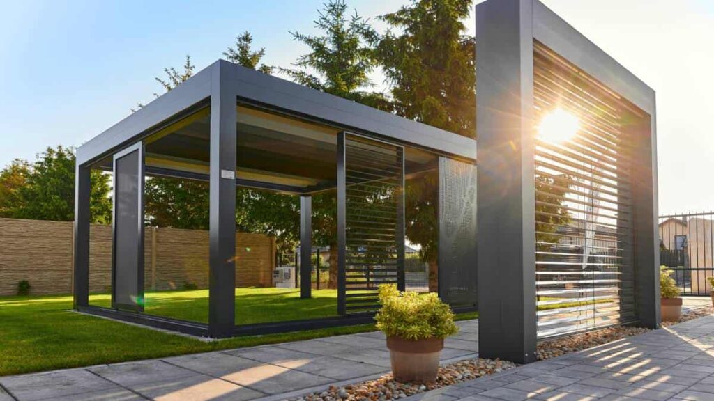 Een zonnige foto van een grijszwarte pergola in het gras naast een terras.